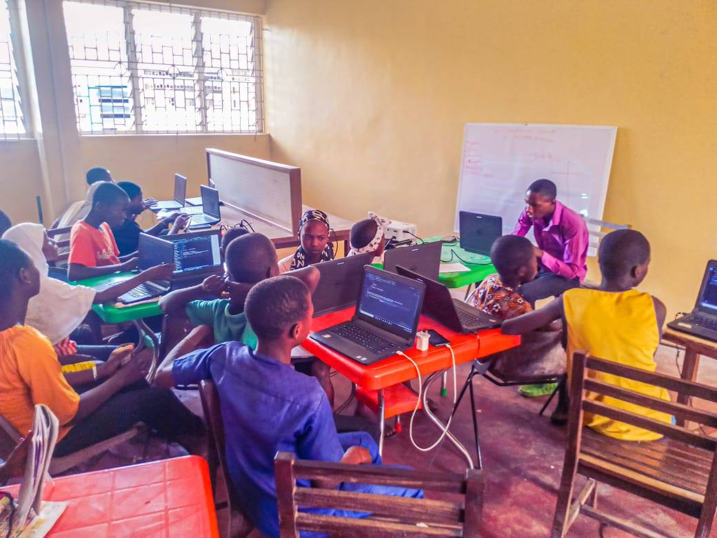 Children working at laptops on brightly coloured desks, with code visible on several screens and an instructor seated at the front.