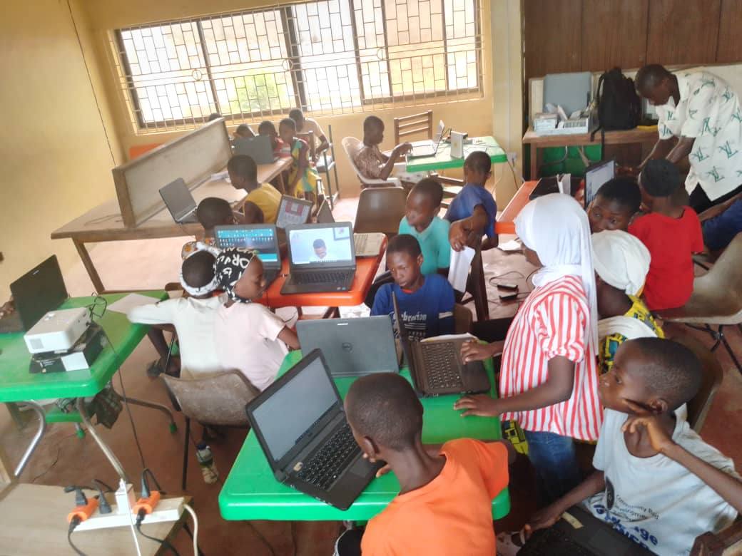 A view across a classroom of children working at laptops set out along green and orange desks.