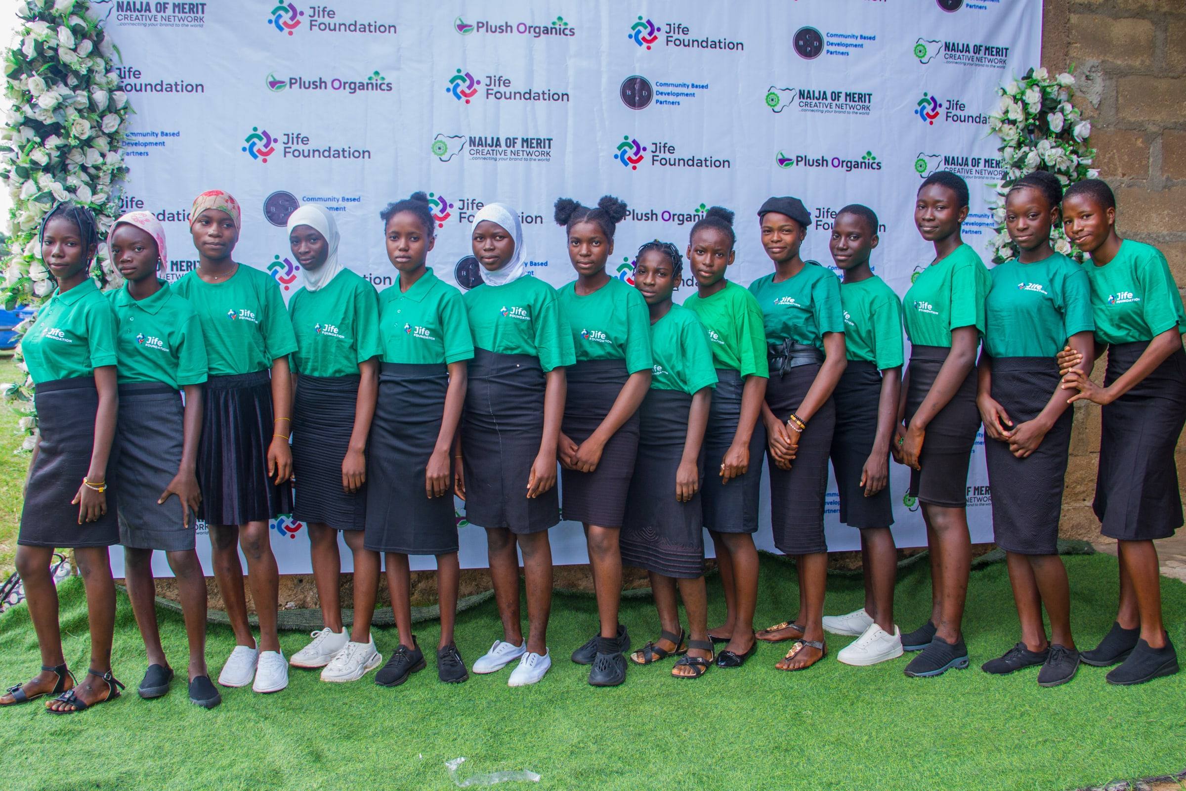 A line of young women in JIFE Foundation T-shirts standing together on grass in front of a JIFE Foundation event backdrop.